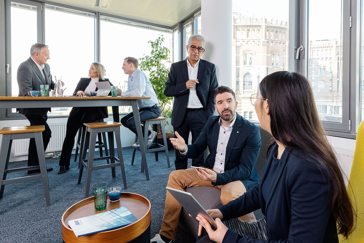 Eine berufliche Besprechung findet in einem hellen Büroraum statt, in dem die Teilnehmer an einem hohen Tisch diskutieren, während andere in der Nähe sitzen.
