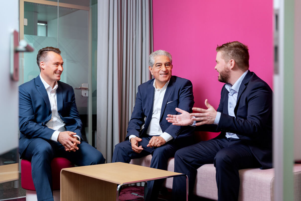 Drei Fachleute in Anzügen diskutieren in einem modernen Büro mit einer leuchtend rosa Wand im Hintergrund.